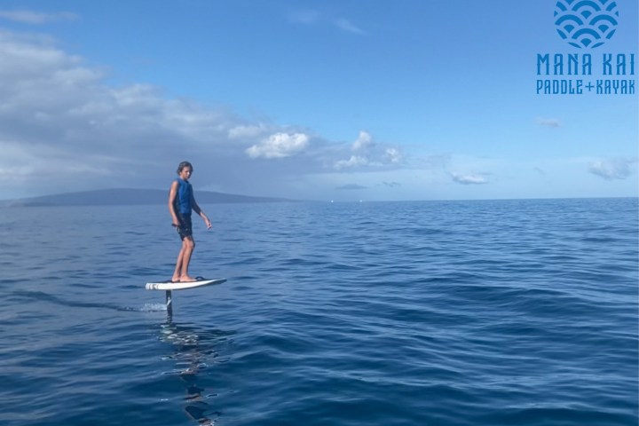 Person riding an electric hydrofoil surfboard on calm ocean waters under a blue sky.